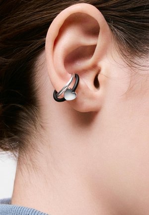 Oreja con dos pendientes: un aro plateado y un aro negro con un colgante de corazón plateado, sobre un primer plano de cabello corto castaño y cuello azul.