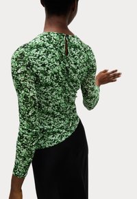 Groene langharige blouse met een textuur en abstract patroon, voorzien van een knoopsluiting aan de achterkant en een getailleerde pasvorm. Zwarte onderkant zichtbaar.