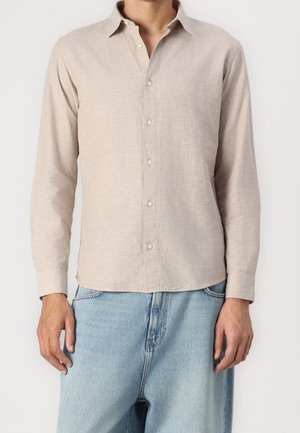 Hombre con camisa beige de manga larga abotonada y vaqueros de mezclilla azul claro de pie frente a un fondo blanco liso.