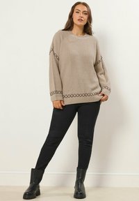 Beige neulepaita, jossa on viistoreunainen pinta ja geometrisiä kirjonnoita hihoissa, yhdistettynä mustiin slim-fit-housuihin ja mustiin nilkkasaappaisiin.