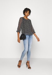 Blusa preta transparente com padrão floral branco, mangas 3/4, combinada com jeans skinny azul claro e sapatos de salto altos pretos com tiras. Pequena bolsa preta.