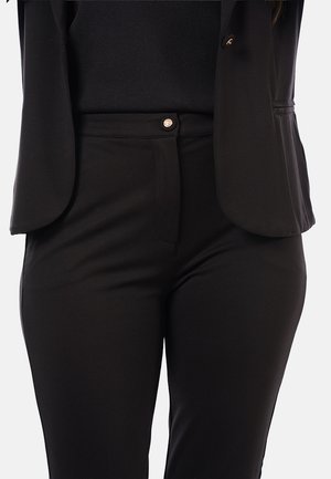Femme portant un pantalon noir ajusté, un haut noir et un blazer noir avec un détail de bouton doré, visible de la mi-torse aux cuisses supérieures.