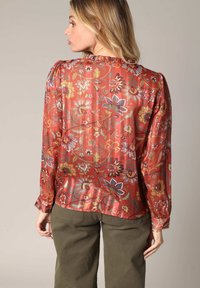 Rode bloemenblouse van een gestructureerde stof, met een gerimpelde halslijn, lange mouwen en een losse pasvorm. De patronen tonen verschillende kleurrijke bloemen.
