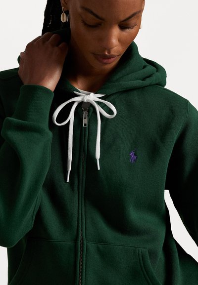 Grøn zip-up hættejakke lavet af blødt stof, med en hvid snøre, frontlynlås og et lille lilla logo på venstre bryst.