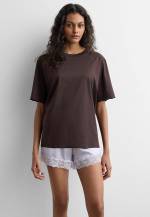 Kobieta o falowanych, ciemnych włosach, ubrana w luźną, ciemnobrązową koszulę z krótkim rękawem i białe szorty wykończone koronką, stojąca na tle jednolitego białego tła.