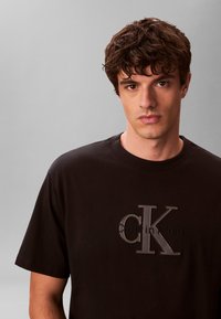 Νέος άνδρας με καστανά σγουρά μαλλιά, φοράει μαύρο T-shirt Calvin Klein, πάνω σε ένα απλό γκρι φόντο.