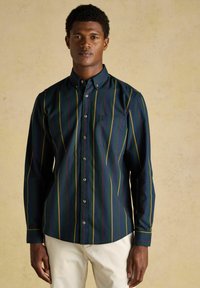 Muž oblečený v tmavozelené košeli na gombíky so žltými a červenými vertikálnymi pruhmi a svetlo béžových nohaviciach, stojaci proti béžovej stene.