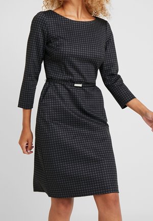 Femme portant une robe cintrée mi-longue noire et gris foncé à motif pied-de-poule, manches trois-quarts et fine ceinture noire.