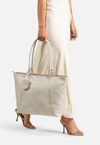 Sac fourre-tout beige avec un motif crocodile, doté de deux poignées et d'une étiquette amovible, associé à des sandales à talons hauts crèmes et nude.