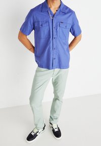 Camisa azul de manga corta con botones y dos bolsillos frontales, combinada con pantalones verde claro y zapatillas negras con detalles blancos y cordones verdes.
