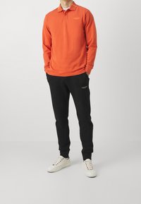 Värikäs oranssi pitkähihainen polo-paita, jossa on kaulus, mustat verkkarit ja valkoiset lenkkarit. Paidassa ja housuissa on pieni logo.