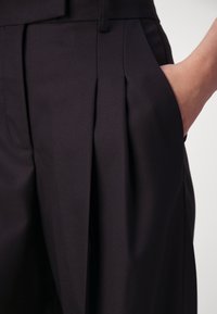 Mustad püksid tekstureeritud kangast, millel on etteplekid ja küljetasku. Disain rõhutab struktureeritud ja sobivat vormi.