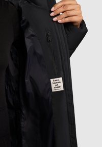 Hand holding open black jacket showing inside pocket with zipper and label reading "A warm hug made out of cloth."