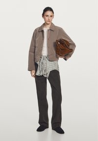 Brown corduroy jacket, white long-sleeve shirt, gray plaid tied around waist, dark pants, holding brown suede bag, black slippers.