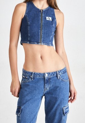Młoda kobieta w bezrękawnym, zapinanym na zamek crop topie z denimu oraz dopasowanych wysokich dżinsowych spodniach cargo z kieszeniami na białym tle.