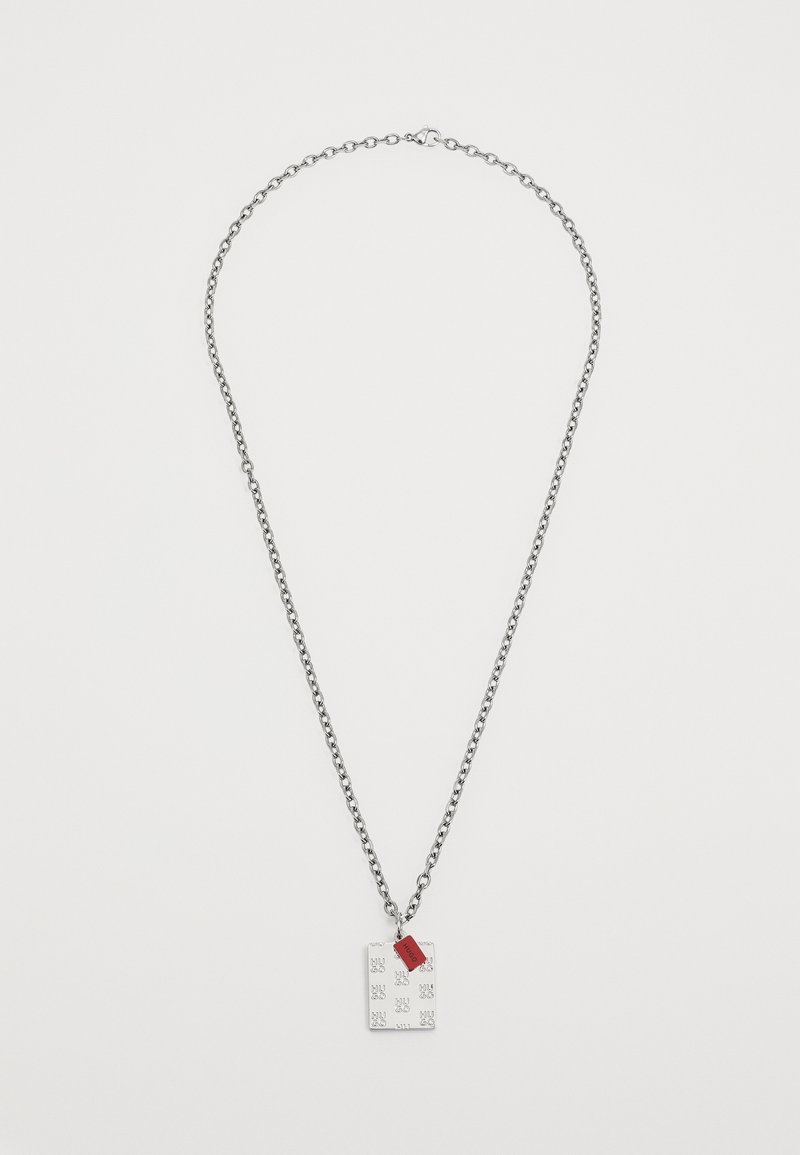 Ασημένια αλυσίδα λαιμού με ορθογώνιο φυλαχτό με ανάγλυφα σχέδια και μια μικρή κόκκινη ετικέτα. Η αλυσίδα έχει υφή.