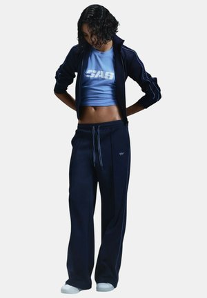 Kobieta w granatowej kurtce i spodniach dresowych z jasnoniebieskim topem, patrzy w dół, stoi na jednolitym tle.