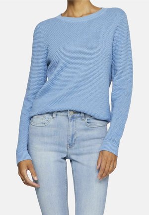 Mujer que lleva un jersey de manga larga azul claro con textura y vaqueros azules claros de tiro alto, con anillos en ambas manos.