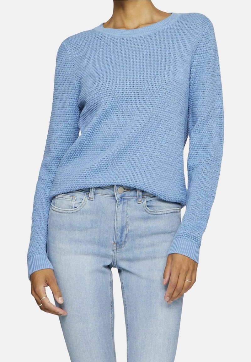 Femme portant un pull texturé bleu clair à manches longues et un jean bleu clair taille haute, avec des bagues aux deux mains.
