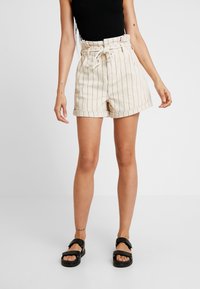 Béžové šortky s vysokým pasem a svislými černými pruhy, se stahováním v pase a ohrnutým lemem, kombinované s černými sandály. Hladká, lehká látka.