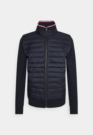 Chaqueta azul marino con panel frontal acolchado, mangas largas de punto, cremallera completa y cuello con botones a presión con ribete rojo y blanco en el interior.