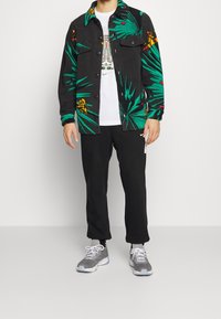 Czarna polarowa kurtka z zielonym wzorem palmowych liści i panterą, noszona na białej koszulce, w połączeniu z czarnymi spodniami i szarymi trampkami.