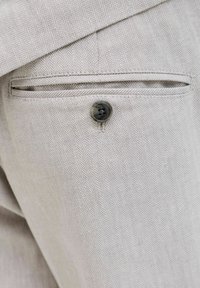 Primo piano di tessuto grigio chiaro a spina di pesce di una tasca posteriore di pantaloni su misura con un bottone nero e apertura a filetto.