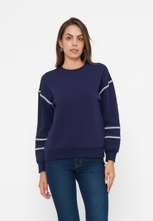 Donna con capelli lunghi e castani che indossa una felpa blu navy con bordo a zigzag bianco sulle maniche e jeans blu, in piedi davanti a uno sfondo bianco.