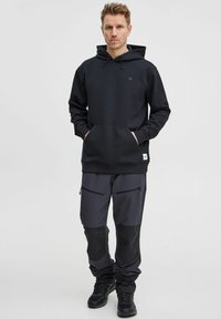 Svart hoodie med framficka och dragsko i huvan, tillsammans med mörkgrå byxor som har zipfickor och kontrasterande knäpaneler.
