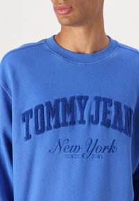 Modra bombažna majica s kapuco z teksturirano vezenino "TOMMY JEANS New York" in "od 1985." Okrugel ovratnik in rebrasti manšeti.