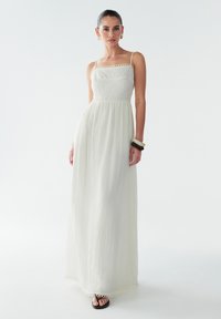 Lange witte jurk met dunne bandjes, gestructureerde stof, nauwsluitend lijfje en uitlopende rok. Afgewerkt met een geschulpte zoom. Accessoires omvatten dikke armbanden.