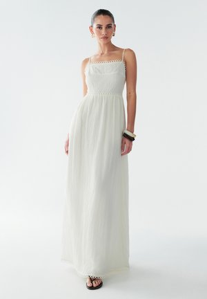 Maxi-kjole i lysecrème stof med en tekstureret overflade, tynde stropper og blondekant ved halsudskæringen og kanten. Sorte sandaler fuldender looket.