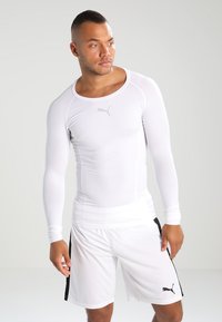 Chemise de sport à manches longues blanche avec un col rond, design ajusté, et logo Puma sur la poitrine. Portée avec un short blanc assorti.