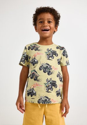 Hymyilevä poika, joka pukeutunut beigekkaan T-paitaan, jossa on monsteriautojen kuvia, ja sinappikeltaisiin shortseihin, seisoo yksivärisen taustan edessä.