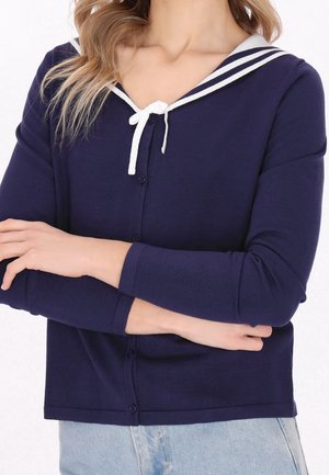 Femme portant un cardigan bleu marine avec col marin blanc et détail nœud, associé à un jean bleu clair, les bras croisés au niveau de la taille.