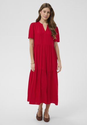 Młoda kobieta o długich włosach, ubrana w czerwoną, warstwową sukienkę typu midi z krótkimi rękawami i brązowe plecione baleriny, stojąca na białym tle.