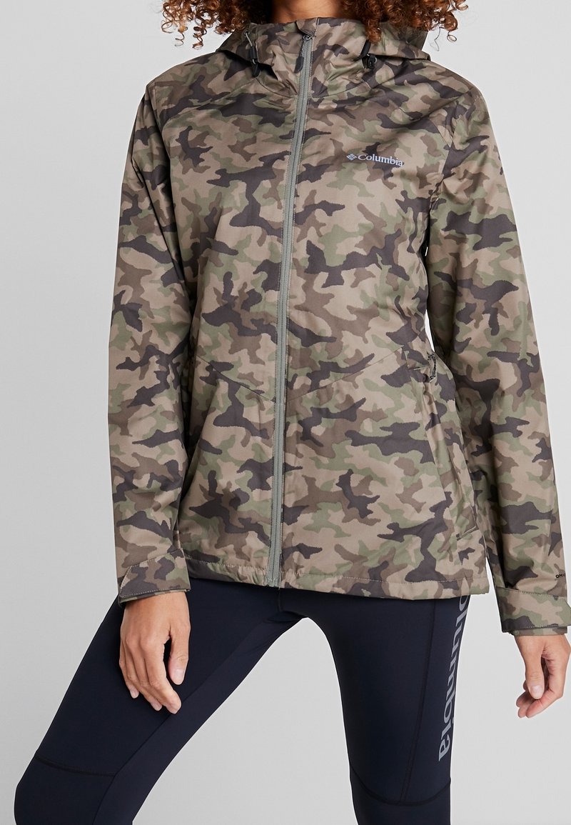 Femme portant une veste à fermeture éclair à capuche camouflage verte et marron, avec un legging noir marqué "Columbia" sur la cuisse.