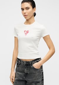 Λευκό βαμβακερό μπλουζάκι με κοντά μανίκια, με ροζ γραφική καρδιά στο κέντρο. Συνδυάζεται με ψηλόμεσα μαύρα τζιν.