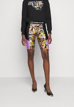 Person trägt schwarzen Hoodie mit floralem Versace Jeans Couture-Logo und schwarze, violette sowie mehrfarbige gemusterte Radlerhosen mit schwarzen High Heels.