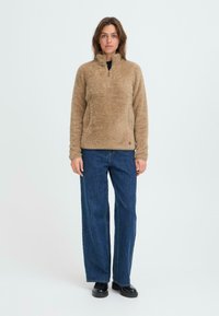 Fleece-Pullover in Beige mit einem Viertel-Reißverschluss und hohem Kragen, kombiniert mit lockeren blauen Jeans und schwarzen Slippern. Weicher, strukturierter Stoff.