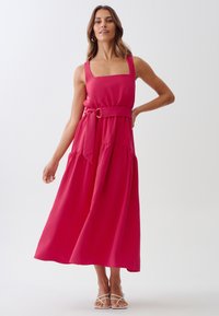 Long pink dress with square neckline, wide straps, and a belted waist. Features a flowing skirt and soft fabric with a slight sheen.