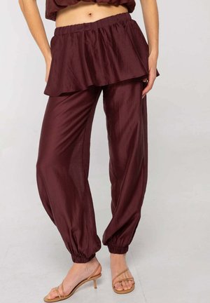 Mujer con pantalones sueltos de color marrón oscuro con tobillos elásticos, combinados con una blusa de varias capas a juego y sandalias de tacón beige con tiras.