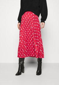 Jupe plissée rouge avec des pois blancs, associée à un pull noir et des bottes noires à talons, arrivant au genou. Texture lisse, longueur mi-mollet.