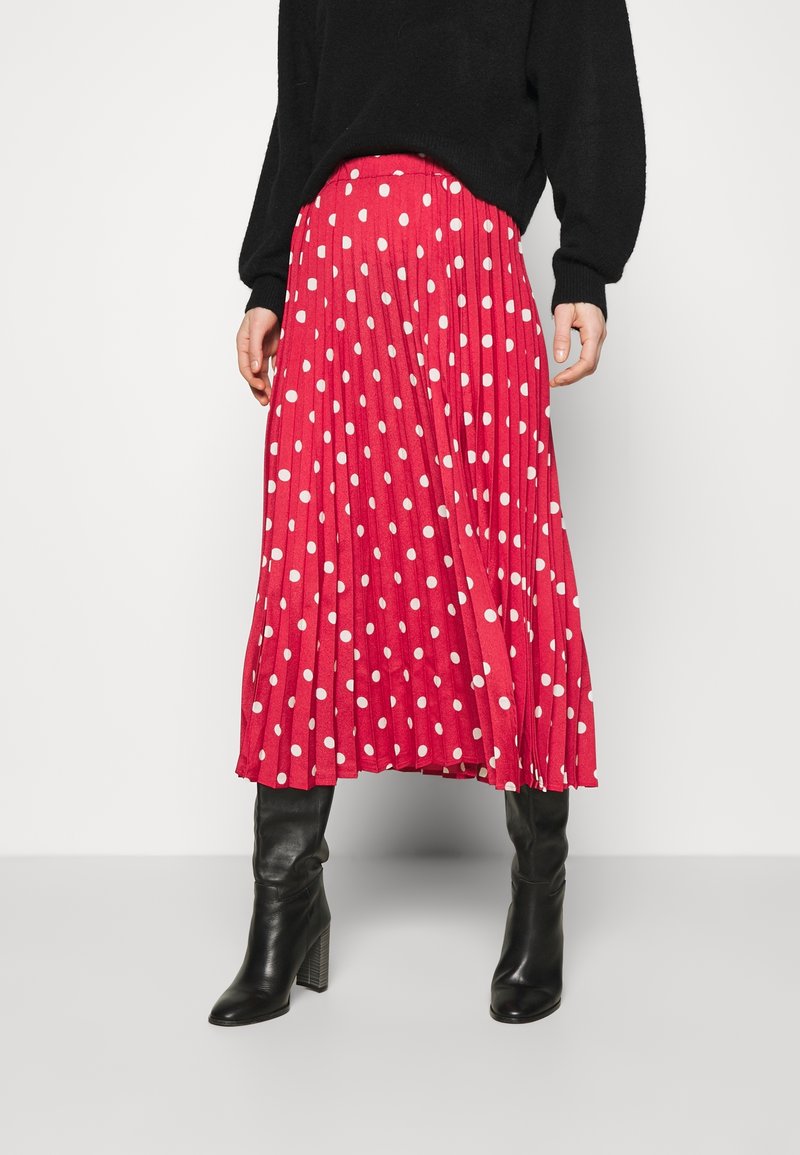 Jupe plissée rouge avec des pois blancs, associée à un pull noir et des bottes noires à talons, arrivant au genou. Texture lisse, longueur mi-mollet.