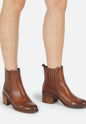 Las botas de cuero marrón hasta el tobillo cuentan con una punta redondeada, paneles laterales texturizados y un tacón de madera grueso, lo que mejora la estabilidad y la comodidad.