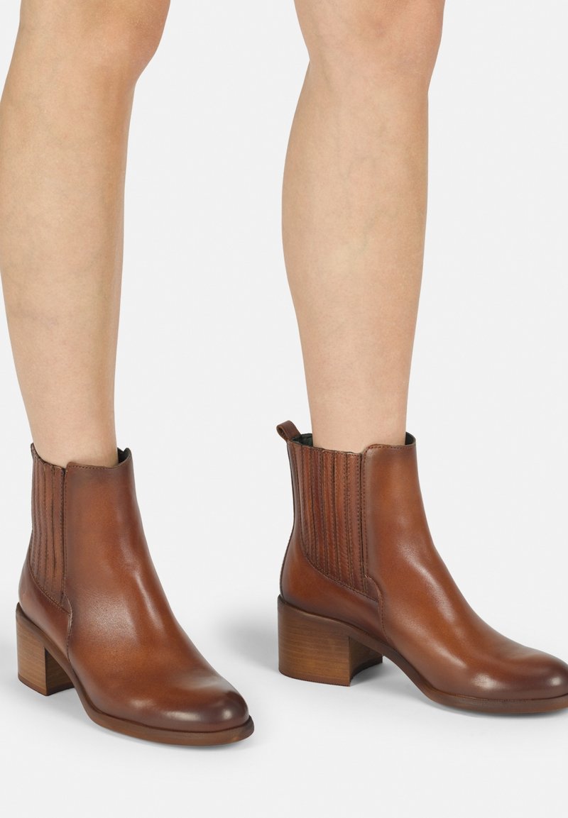 Las botas de cuero marrón hasta el tobillo cuentan con una punta redondeada, paneles laterales texturizados y un tacón de madera grueso, lo que mejora la estabilidad y la comodidad.