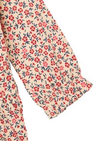 Lichtbeige stof met een bloemmotief met rode en marineblauwe bloemen. De lange mouwmanchet is geplooid, wat textuur en detail toevoegt.