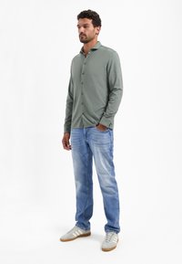 Langarm, knoop-up shirt in salie groen, gecombineerd met lichtblauwe jeans. Het shirt heeft een kraag en een subtiele textuur; de schoenen zijn grijs met witte strepen.