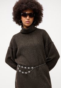 Maglione marrone oversize con collo a imbuto. Cintura in catena argentata decorata con forme a stella. Modella che indossa occhiali da sole neri e orecchini a cerchio.