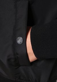 Wnętrze rękawa czarnej kurtki z gładką tkaniną i ściągaczem z dzianiny. W pobliżu kieszeni widoczny jest srebrny metalowy zatrzask.